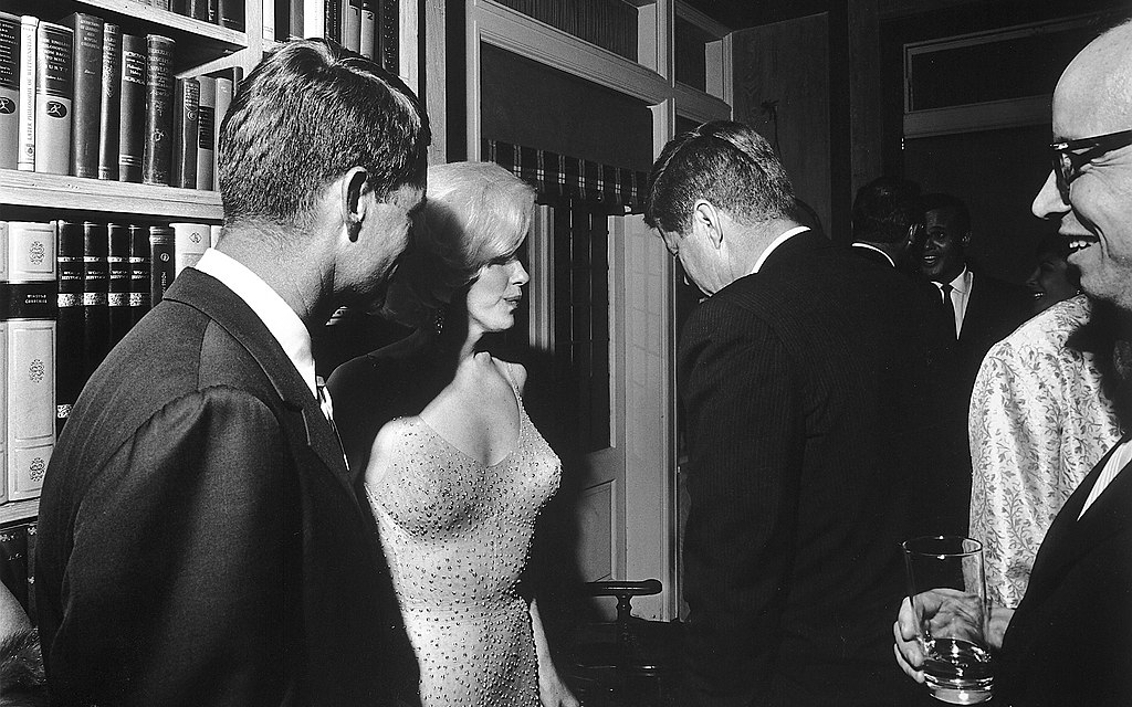 U.S. President John F. Kennedy (with his back to the camera), U.S. Attorney General Robert Kennedy (far left), and actress Marilyn Monroe, on the occasion of President Kennedy's 45th birthday celebrations at Madison Square Garden in New York City. Arthur M. Schlesinger, Jr. is at the far right. Facing the camera in the rear appears singer Harry Belafonte. 19 May 1962. Cecil W. Stoughton, official White House photographer. This image is in the public domain. Image location on Wikimedia Commons.